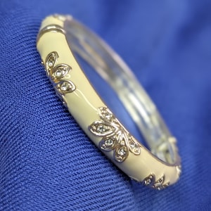 May include: Cream-colored bangle bracelet with silver-tone floral accents and clear crystal embellishments. The bracelet has a hinged closure and is set against a blue background. A stylish piece of jewelry.