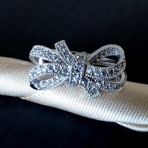 May include: A silver bow ring, adorned with sparkling gems. The ring sits on a cream-coloured fabric, set against a dark backdrop. The bow design is intricate and visually appealing, ideal for adding a touch of elegance.
