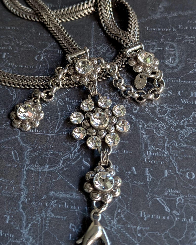 Peut inclure: Collier argent&eacute; orn&eacute; de plusieurs pendentifs en forme de fleurs, chacun orn&eacute; de cristaux clairs et &eacute;tincelants. La cha&icirc;ne a un design tiss&eacute;. Le collier est pr&eacute;sent&eacute; sur un fond sombre avec un motif de carte.
