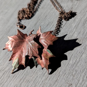 Puede incluir: Un collar de color cobre con dos colgantes detallados de hojas de arce. Las hojas tienen una superficie texturizada con toques verdes y están unidas a una delicada cadena. El collar se exhibe sobre una superficie de madera gris.