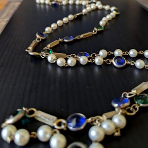May include: A vintage necklace featuring faux pearls, gold-tone metal accents, and jewel-toned blue and green stones. The necklace is displayed on a dark surface, showcasing its intricate design and classic style. The necklace is a beautiful piece of jewelry.