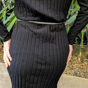 Peut inclure: Robe noire en maille c&ocirc;tel&eacute;e &agrave; manches longues et coupe ajust&eacute;e. La robe est cintr&eacute;e &agrave; la taille par une fine ceinture argent&eacute;e. Le tissu pr&eacute;sente des lignes verticales, cr&eacute;ant un aspect textur&eacute;.