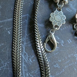 Peut inclure: Gros plan sur un collier argent&eacute; avec un motif de cha&icirc;ne tress&eacute;e. Le collier comporte des pendentifs floraux d&eacute;coratifs et un fermoir mousqueton. Le fond est une carte.