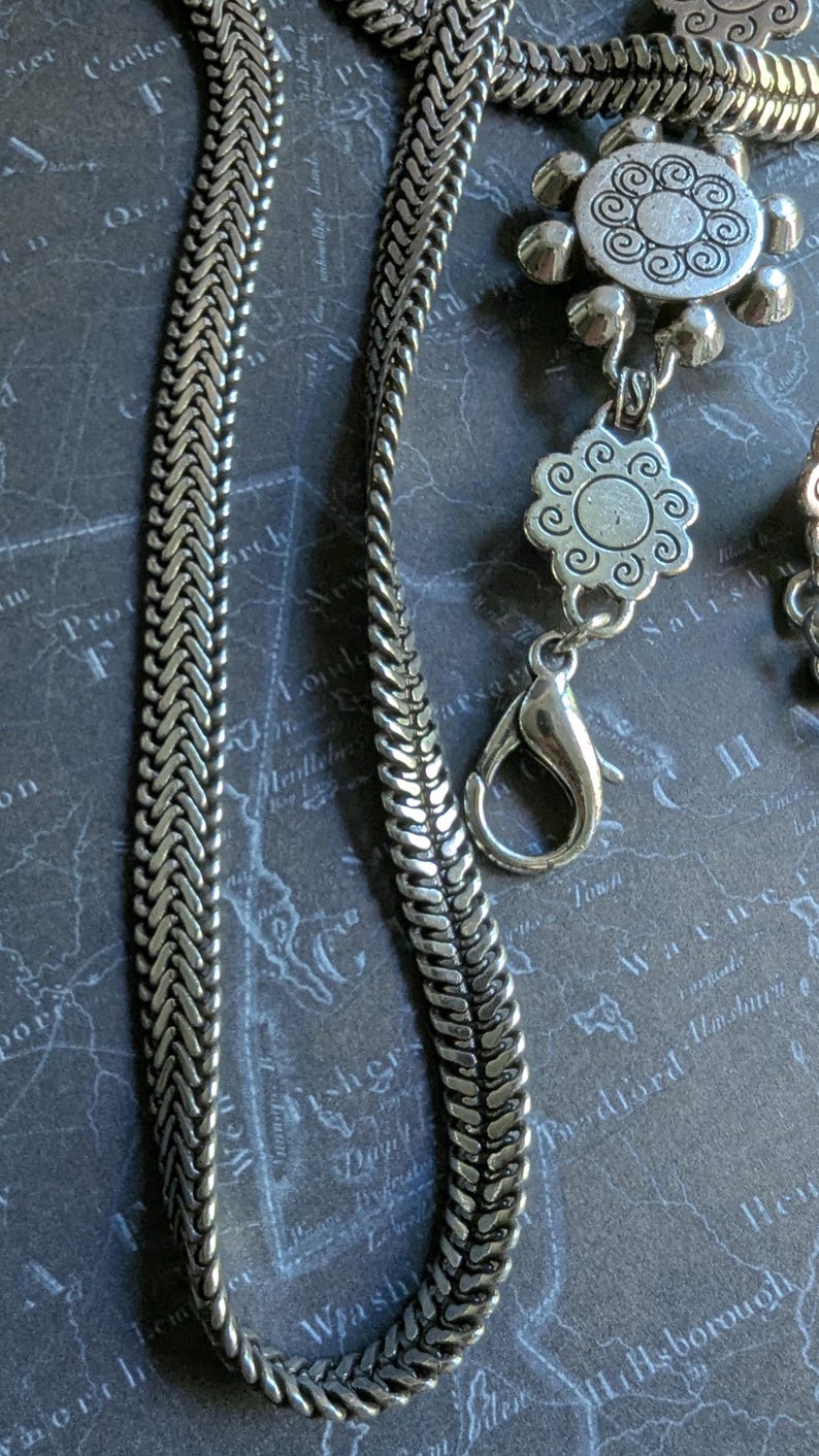 Peut inclure: Gros plan sur un collier argent&eacute; avec un motif de cha&icirc;ne tress&eacute;e. Le collier comporte des pendentifs floraux d&eacute;coratifs et un fermoir mousqueton. Le fond est une carte.