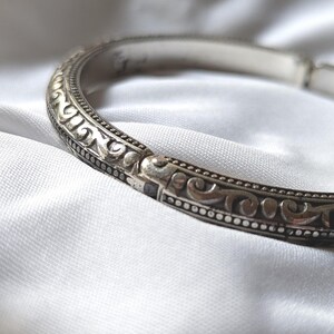 May include: A silver-toned bracelet with an ornate, repeating scroll design. The bracelet is set against a white, textured background. The design features a series of small, raised dots along the edges.