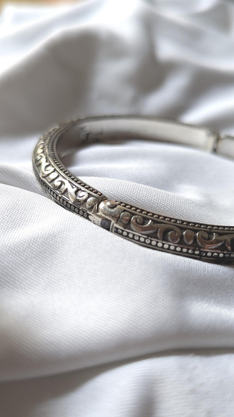 May include: A silver-toned bracelet with an ornate, repeating scroll design. The bracelet is set against a white, textured background. The design features a series of small, raised dots along the edges.