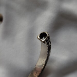 May include: Silver-toned cuff bracelet with an open design. The bracelet features a detailed, textured pattern along the edges and a decorative end piece with a dark, hollowed-out design. The bracelet is shown against a soft, neutral background.