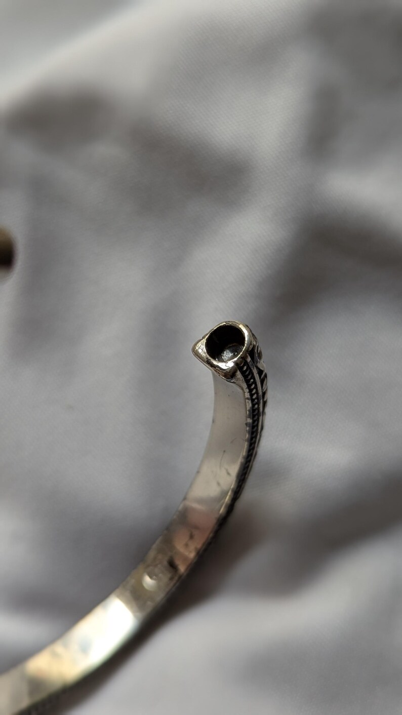 May include: Silver-toned cuff bracelet with an open design. The bracelet features a detailed, textured pattern along the edges and a decorative end piece with a dark, hollowed-out design. The bracelet is shown against a soft, neutral background.