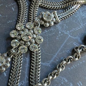 Peut inclure: Collier argent&eacute; avec diff&eacute;rents styles de cha&icirc;nes, dont un motif tress&eacute; et une cha&icirc;ne d&eacute;licate. Le collier comporte plusieurs pendentifs en forme de fleur orn&eacute;s de cristaux clairs et &eacute;tincelants. Le bijou est pr&eacute;sent&eacute; sur un fond sombre.