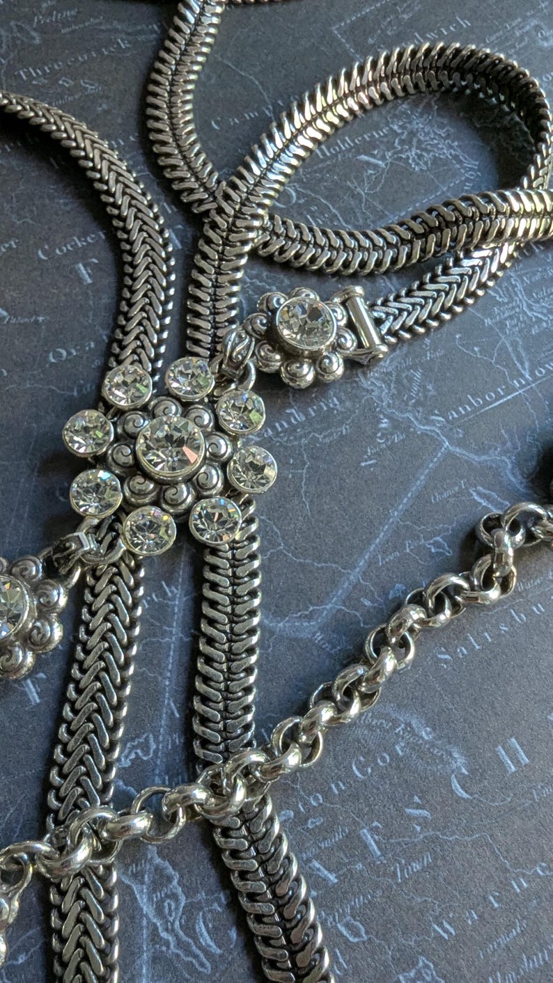 Peut inclure: Collier argent&eacute; avec diff&eacute;rents styles de cha&icirc;nes, dont un motif tress&eacute; et une cha&icirc;ne d&eacute;licate. Le collier comporte plusieurs pendentifs en forme de fleur orn&eacute;s de cristaux clairs et &eacute;tincelants. Le bijou est pr&eacute;sent&eacute; sur un fond sombre.