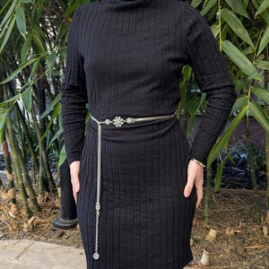 Peut inclure: Une robe noire &agrave; col roul&eacute; c&ocirc;tel&eacute;e avec des manches longues. Une ceinture cha&icirc;ne argent&eacute;e avec un motif floral et une cha&icirc;ne pendante cintre la taille. La robe est port&eacute;e devant un bambou.