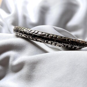 May include: Silver-toned bangle bracelet with intricate, repeating floral and scrollwork patterns. The bracelet is set against a white, textured fabric background, highlighting the detailed design and metallic sheen of the jewelry.
