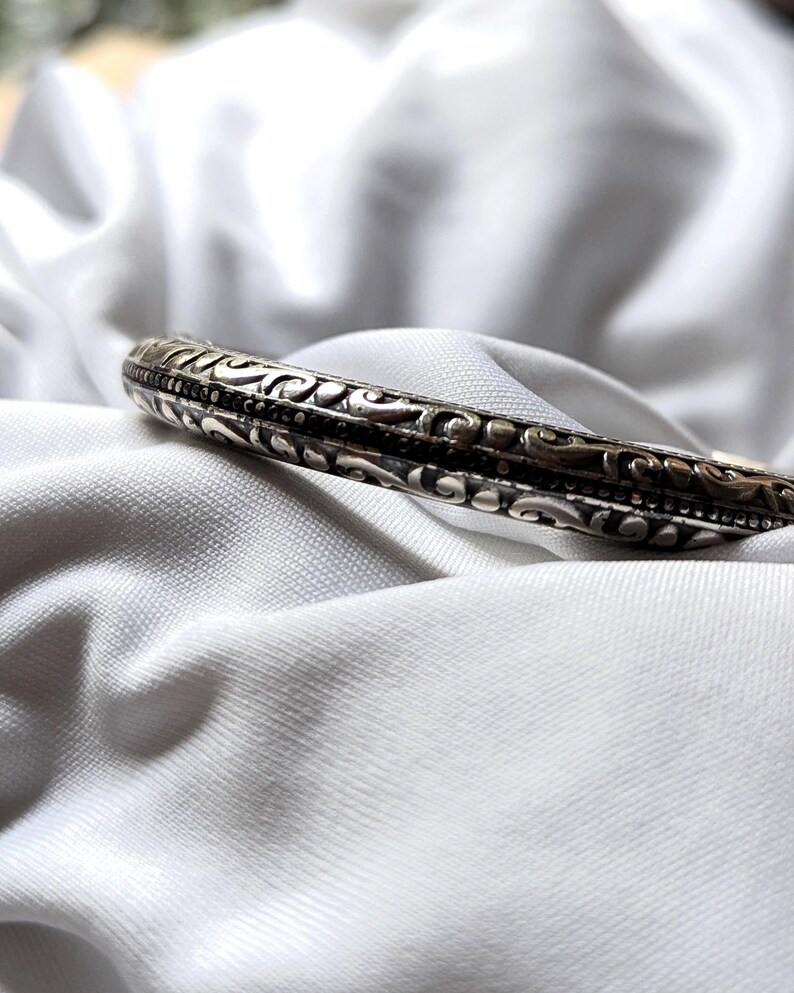 May include: Silver-toned bangle bracelet with intricate, repeating floral and scrollwork patterns. The bracelet is set against a white, textured fabric background, highlighting the detailed design and metallic sheen of the jewelry.