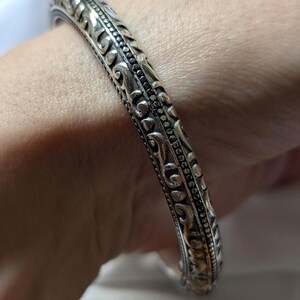 May include: Ornate silver bangle bracelet with detailed scrollwork and a textured design. The bracelet is a solid band with a repeating pattern of swirling lines and curves. The metal has a polished finish.
