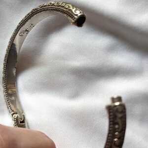 May include: A silver-colored hinged bangle bracelet with an ornate, engraved design. The bracelet features a detailed pattern of swirls and dots along its surface. The clasp is visible, and the bracelet is set against a white fabric background.