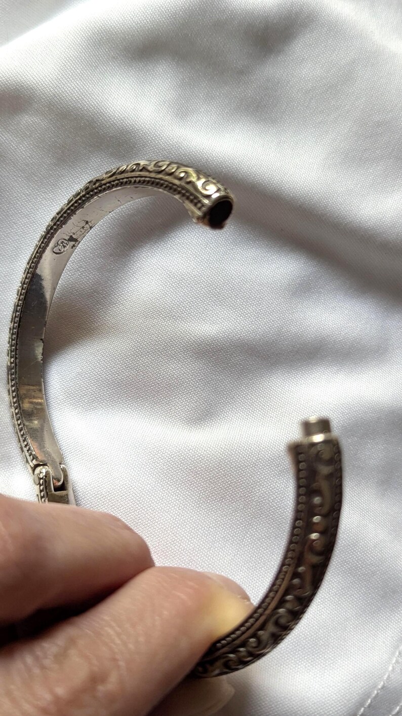May include: A silver-colored hinged bangle bracelet with an ornate, engraved design. The bracelet features a detailed pattern of swirls and dots along its surface. The clasp is visible, and the bracelet is set against a white fabric background.