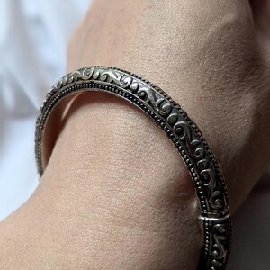 May include: Silver-toned bangle bracelet with intricate scrollwork and beaded detailing. The hinged bracelet is shown on a person's wrist, showcasing the ornate design and secure clasp. A classic piece of jewelry.