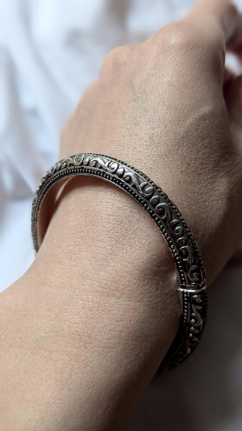 May include: Silver-toned bangle bracelet with intricate scrollwork and beaded detailing. The hinged bracelet is shown on a person's wrist, showcasing the ornate design and secure clasp. A classic piece of jewelry.