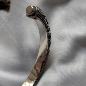 May include: Silver-toned cuff bracelet with an open design. The bracelet features intricate, dark-toned detailing along the edges and at the ends. The metal has a polished, reflective surface.