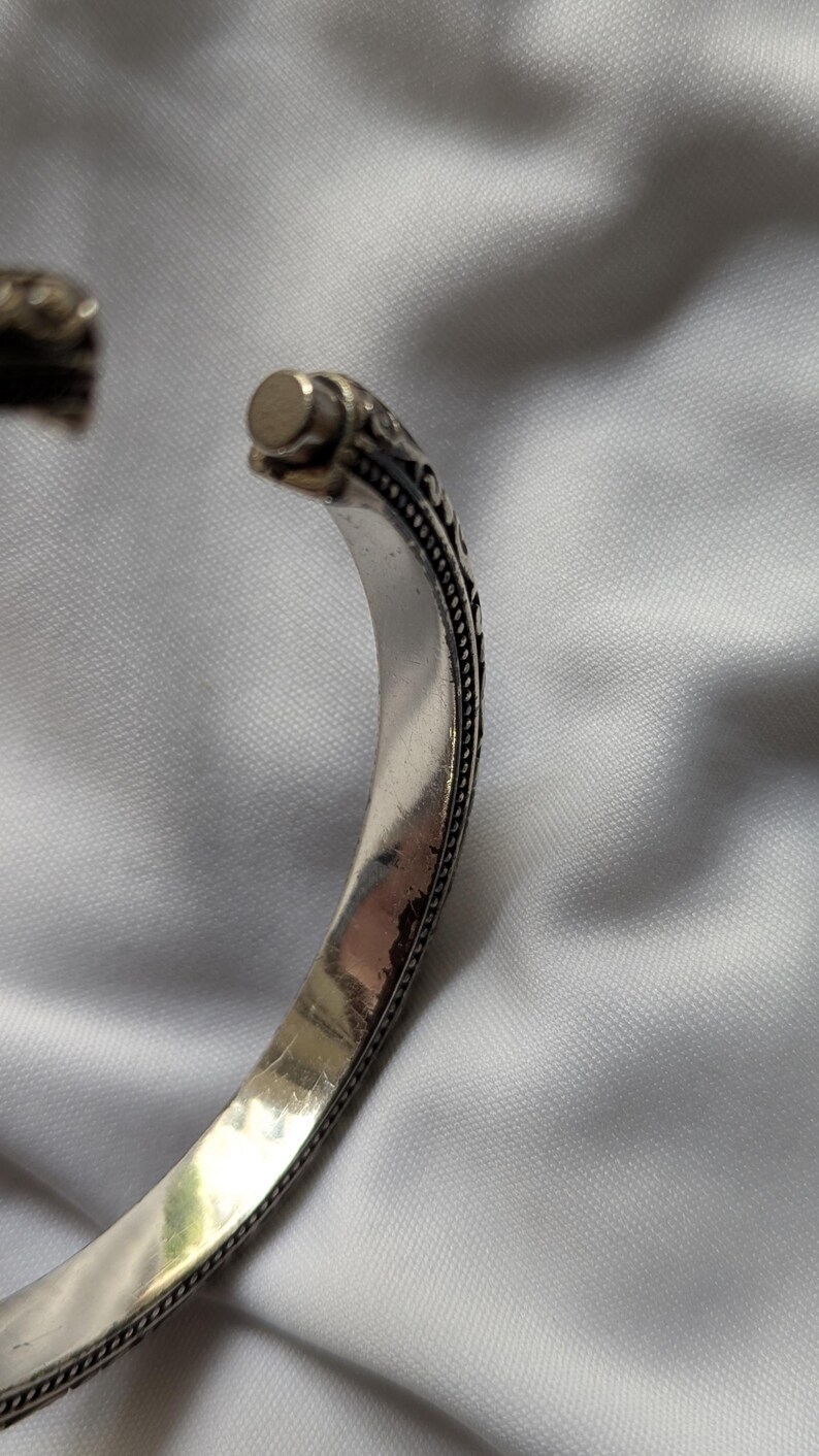 May include: Silver-toned cuff bracelet with an open design. The bracelet features intricate, dark-toned detailing along the edges and at the ends. The metal has a polished, reflective surface.