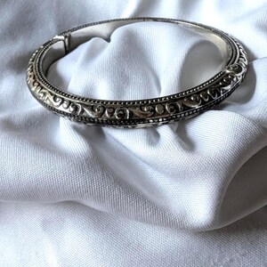 May include: A silver hinged bangle bracelet with intricate scrollwork and beaded detailing. The bracelet is displayed on a white, textured fabric, showcasing its ornate design and craftsmanship. The bracelet's clasp is visible.