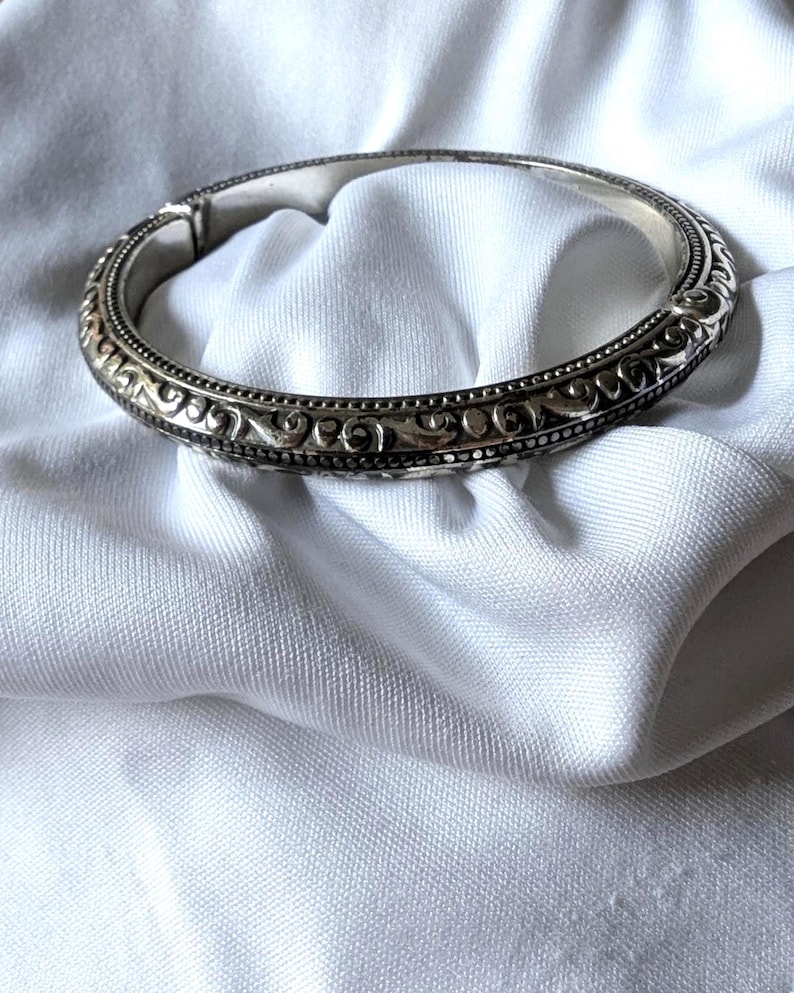 May include: A silver hinged bangle bracelet with intricate scrollwork and beaded detailing. The bracelet is displayed on a white, textured fabric, showcasing its ornate design and craftsmanship. The bracelet's clasp is visible.