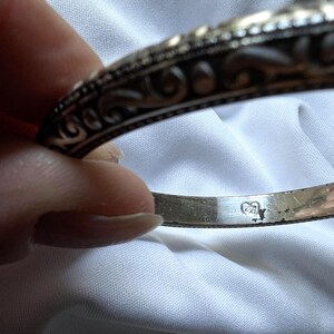 May include: Close-up of a silver bangle bracelet with ornate scrollwork and a hallmark stamp. The bracelet has a detailed, textured design and a polished finish. The background is a white fabric.