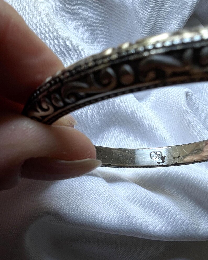 May include: Close-up of a silver bangle bracelet with ornate scrollwork and a hallmark stamp. The bracelet has a detailed, textured design and a polished finish. The background is a white fabric.
