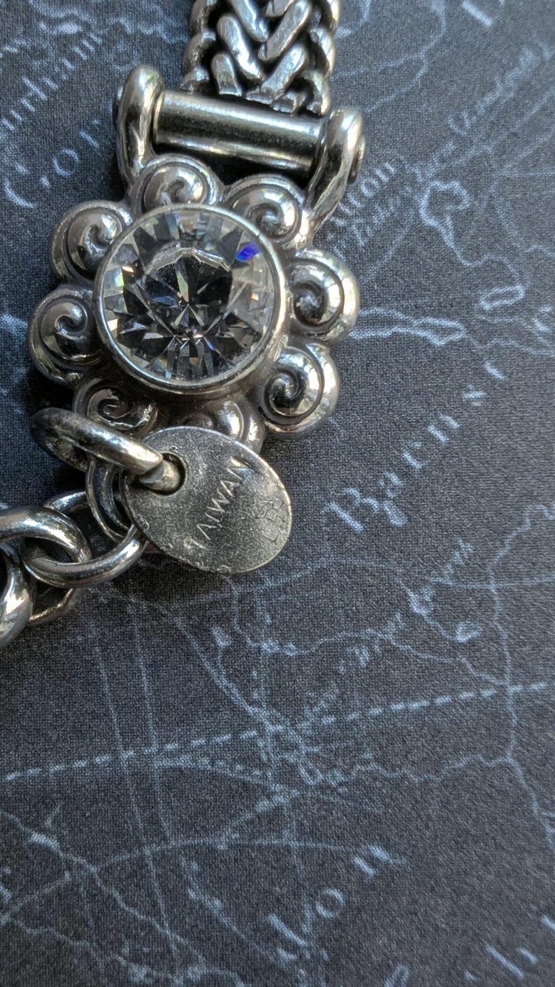 Peut inclure: Gros plan sur un bracelet argent&eacute; avec une grosse pierre pr&eacute;cieuse ronde et transparente sertie dans un motif floral. Le bracelet a une cha&icirc;ne et une petite &eacute;tiquette avec l'inscription "TAIWAN". Le fond est une surface gris fonc&eacute; avec un motif de carte.