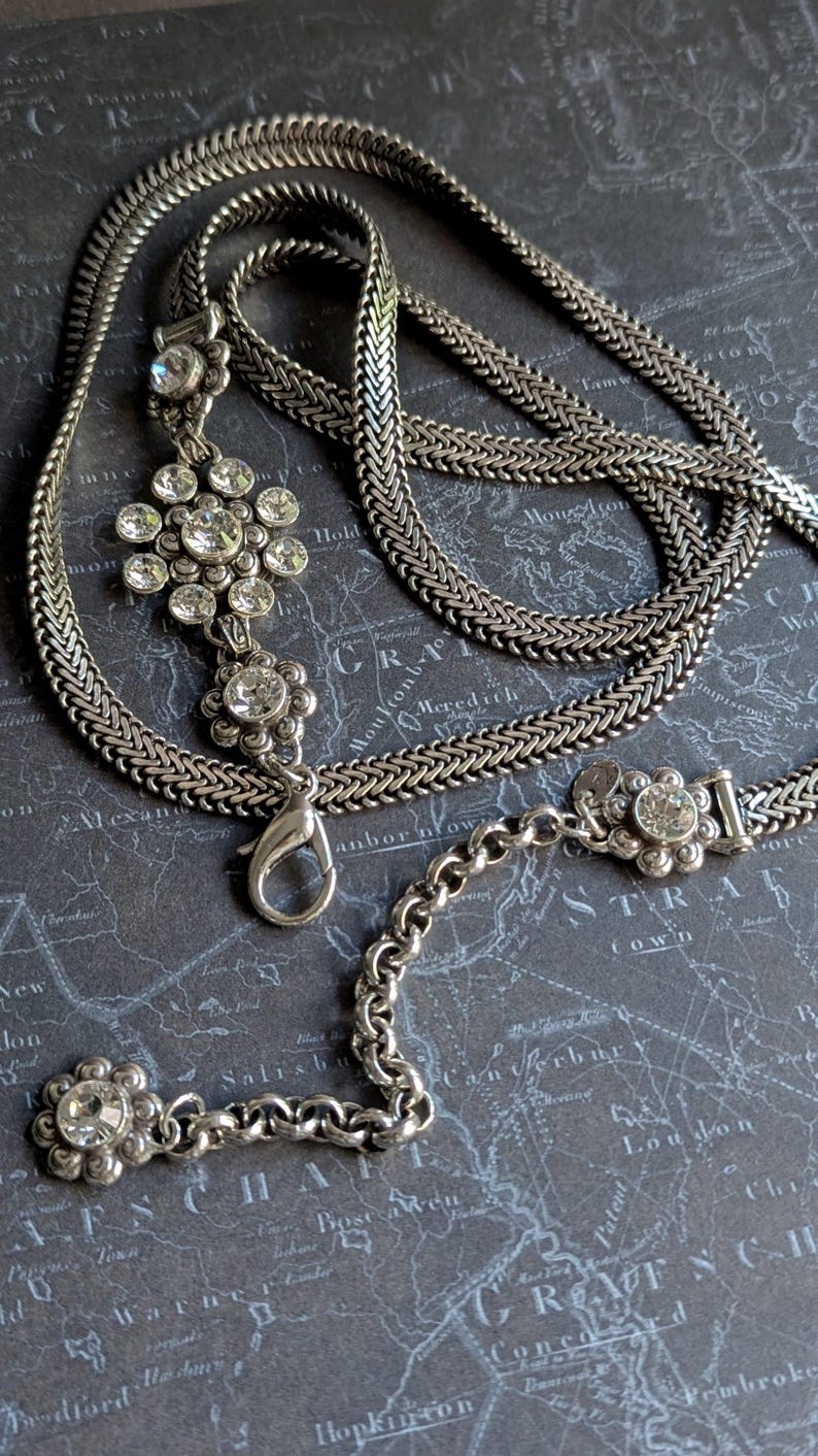 Peut inclure: Collier argent&eacute; avec un pendentif floral et des cristaux transparents &eacute;tincelants. Le collier pr&eacute;sente un motif de cha&icirc;ne et un fermoir mousqueton. Le pendentif est en forme de fleur avec plusieurs cristaux. Le collier est pr&eacute;sent&eacute; sur un fond gris fonc&eacute;.