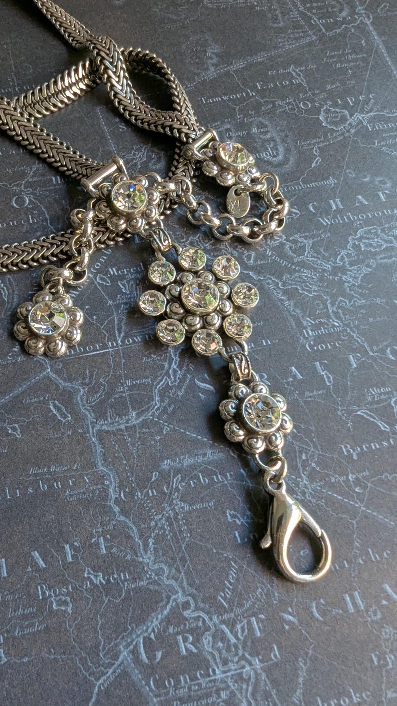 Peut inclure: Collier argent&eacute; orn&eacute; de pendentifs en forme de fleurs et de cristaux transparents. Le collier pr&eacute;sente un motif de cha&icirc;ne et une fermeture &agrave; mousqueton. Le bijou est pr&eacute;sent&eacute; sur une surface bleu fonc&eacute; &agrave; motif de carte.