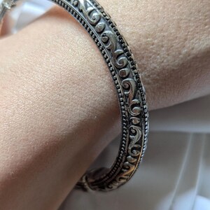 May include: Silver-toned bangle bracelet with an ornate, repeating scroll design. The bracelet has a detailed pattern of swirling lines and small dots. The jewelry is a classic accessory.