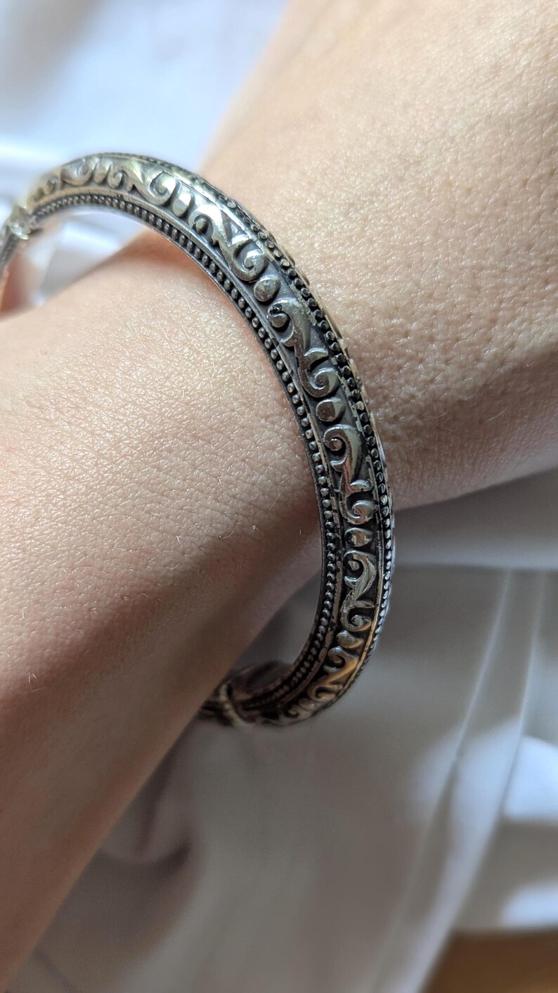 May include: Silver-toned bangle bracelet with an ornate, repeating scroll design. The bracelet has a detailed pattern of swirling lines and small dots. The jewelry is a classic accessory.