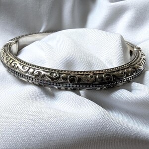May include: Silver-toned bangle bracelet with an ornate, repeating pattern of swirls and dots. The bracelet is set against a white fabric background, highlighting its intricate design and metallic sheen. A classic piece of jewelry.