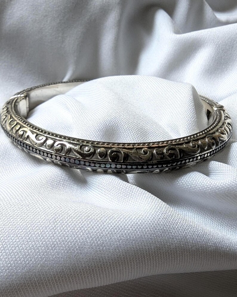 May include: Silver-toned bangle bracelet with an ornate, repeating pattern of swirls and dots. The bracelet is set against a white fabric background, highlighting its intricate design and metallic sheen. A classic piece of jewelry.