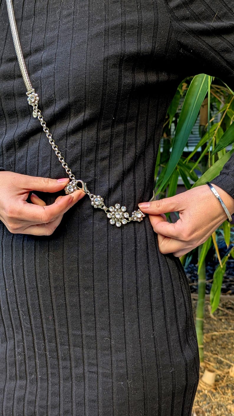 Peut inclure: Ceinture argent&eacute;e orn&eacute;e de strass floraux. La ceinture est attach&eacute;e autour d'une robe noire c&ocirc;tel&eacute;e. Le design de la ceinture pr&eacute;sente une cha&icirc;ne et une s&eacute;rie de strass clairs et &eacute;tincelants dispos&eacute;s en motif floral.