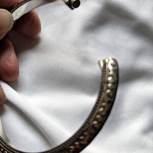 May include: A silver-colored cuff bracelet with intricate embossed patterns. The open bracelet is held against a white fabric background. The bracelet has a detailed design along the edges.