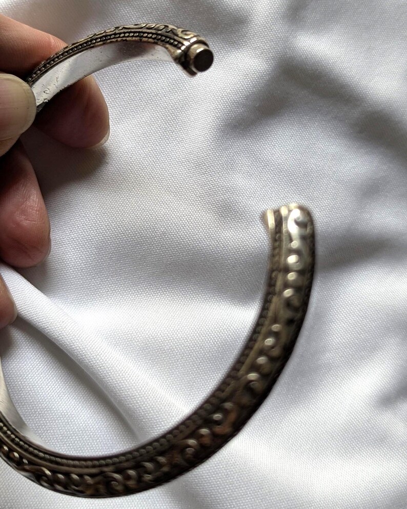May include: A silver-colored cuff bracelet with intricate embossed patterns. The open bracelet is held against a white fabric background. The bracelet has a detailed design along the edges.