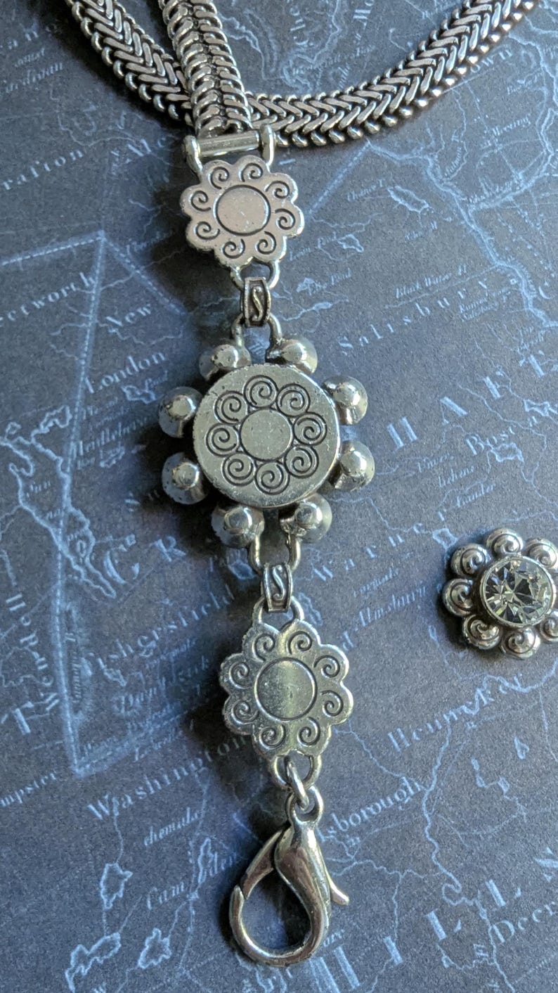 Peut inclure: Collier argent&eacute; avec des motifs de pendentifs floraux et circulaires. Le collier est dot&eacute; d'un fermoir mousqueton et d'une petite boucle d'oreille assortie en forme de fleur avec un centre en cristal transparent. Le bijou est pos&eacute; sur un fond de carte.