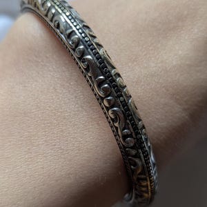 May include: Silver-toned bangle bracelet with intricate, embossed floral and scrollwork patterns. The bracelet has a slightly curved shape and a detailed design that wraps around the entire band. A classic piece of jewelry.