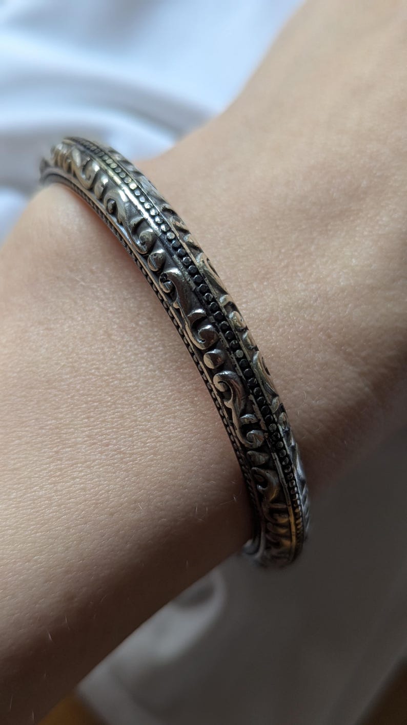 May include: Silver-toned bangle bracelet with intricate, embossed floral and scrollwork patterns. The bracelet has a slightly curved shape and a detailed design that wraps around the entire band. A classic piece of jewelry.