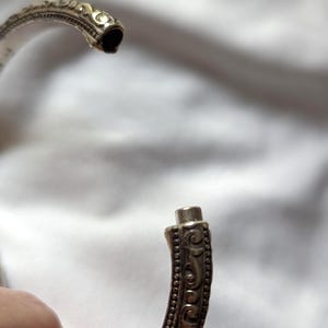 May include: Close-up of an open silver-colored bracelet with intricate, ornate detailing. The bracelet's design features a series of small, raised dots and swirling patterns. The clasp is visible, showing how the bracelet opens and closes.