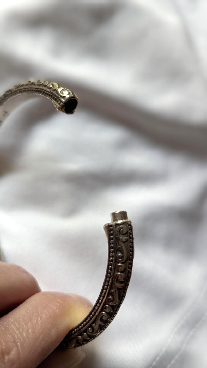 May include: Close-up of an open silver-colored bracelet with intricate, ornate detailing. The bracelet's design features a series of small, raised dots and swirling patterns. The clasp is visible, showing how the bracelet opens and closes.