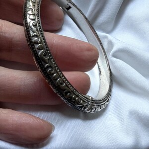 May include: A silver hinged bangle bracelet with ornate scrollwork and a textured border. The bracelet is held against a white background, showcasing its intricate design and polished finish. The clasp is visible.