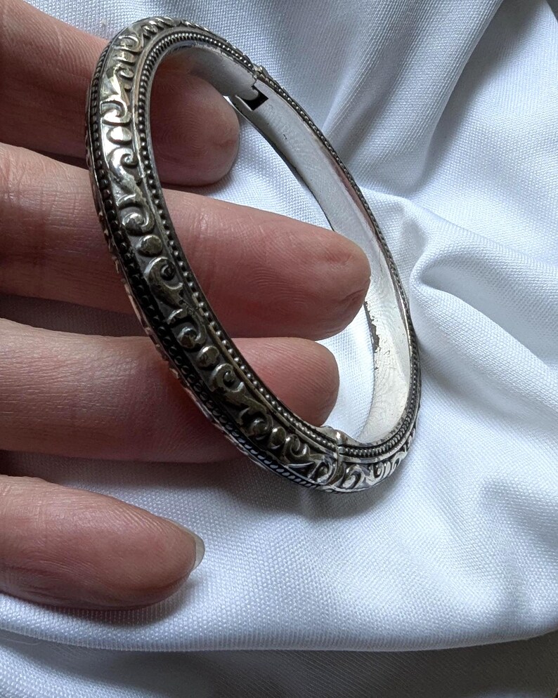 May include: A silver hinged bangle bracelet with ornate scrollwork and a textured border. The bracelet is held against a white background, showcasing its intricate design and polished finish. The clasp is visible.
