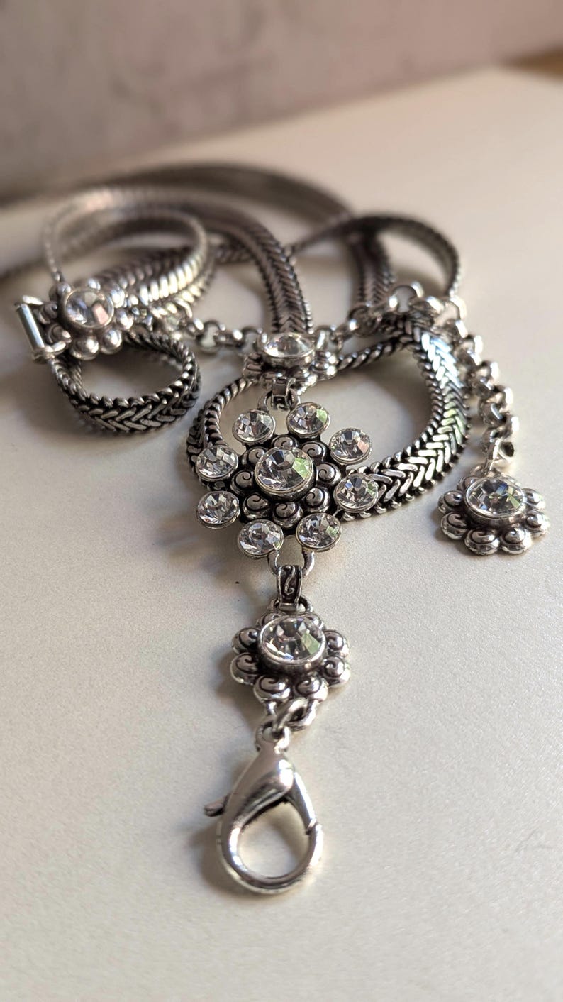 Peut inclure: Un collier argent&eacute; avec un pendentif floral et des cristaux transparents &eacute;tincelants. Le collier pr&eacute;sente une cha&icirc;ne tress&eacute;e et un bracelet assorti avec un fermoir floral. Le pendentif a un motif floral avec plusieurs cristaux.