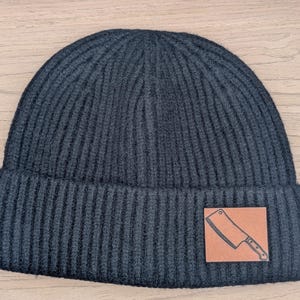 May include: Dark gray ribbed knit beanie with a brown leather patch featuring a cleaver graphic. The hat is a classic, warm winter accessory.