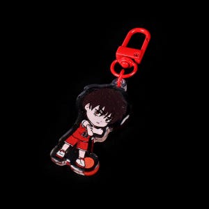 May include: Red and black anime character keychain with a basketball. The character is a boy with brown hair and a red jersey. The keychain has a red metal clasp.