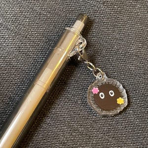 May include: A clear, silver pen with a small charm attached. The charm is a brown circle with two eyes and two small flowers, one pink and one yellow. The pen is on a gray surface.