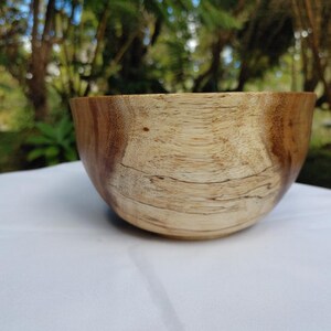 May include: A wooden bowl with a natural finish. The bowl has a light brown interior and a darker brown exterior. The wood grain is visible. The bowl is sitting on a white surface.